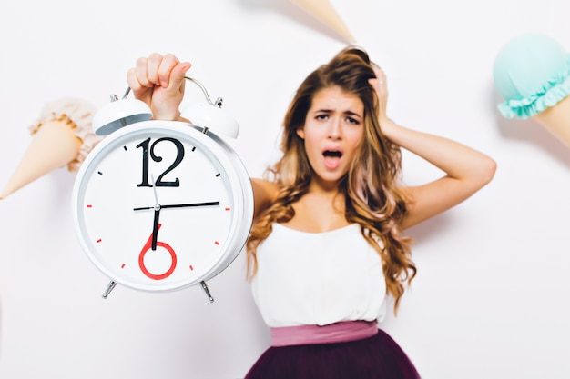 Retrato de primer plano de mujer joven infeliz con cabello largo brillante, tocando su cabeza en pánico. Chica desafortunada con ropa de moda sosteniendo un gran reloj llega tarde al trabajo y grita.