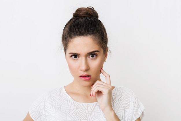 Retrato de primer plano de mujer joven pensante en blusa blanca mordiéndose el labio, frunciendo el ceño, sosteniendo el dedo en su cara, expresión facial seria y emocional, teniendo un problema aislado