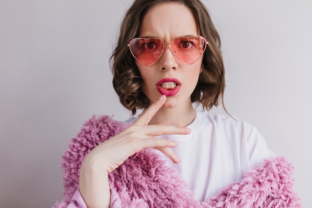 Retrato de primer plano de una mujer morena descontenta con gafas de sol haciendo muecas en la pared blanca. Filmación en interiores de moda chica caucásica en abrigo de piel rosa.