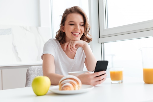 Retrato de primer plano de mujer morena feliz usando teléfono móvil mientras desayunando en la cocina blanca