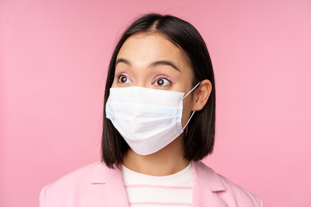 Retrato de primer plano de una mujer de negocios asiática con mascarilla médica que parece sorprendida de pie en traje sobre fondo rosa