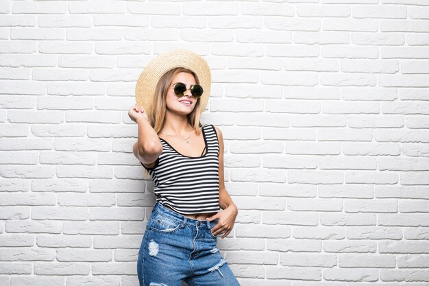 Retrato de primer plano de mujer posando moda sombrero de paja y gafas de sol contra la pared de ladrillo blanco.