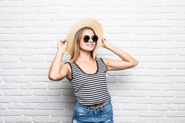 Retrato de primer plano de mujer posando moda sombrero de paja y gafas de sol contra la pared de ladrillo blanco.