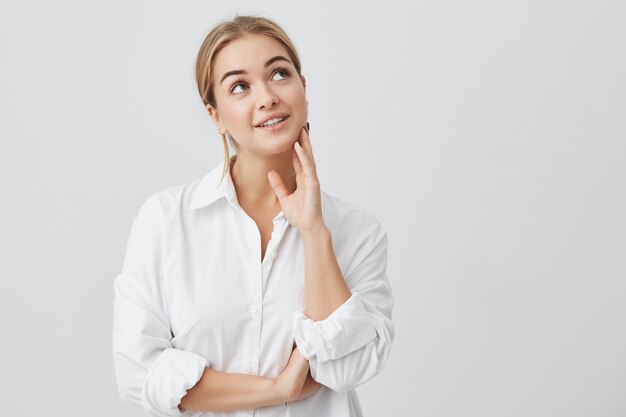 Retrato de primer plano de mujer satisfecha con piel pura, ojos oscuros y sonrisa sincera con camisa blanca posando. Mujer soñadora pensando en cosas agradables, mirando hacia arriba.