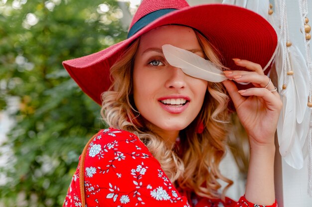 Retrato de primer plano de mujer sonriente rubia con estilo atractivo en traje de moda de verano de sombrero rojo de paja y blusa con piel de cara sensual sexy de plumas blancas