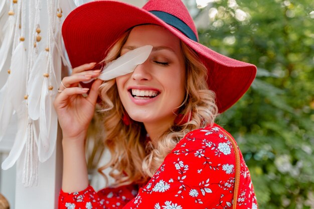 Retrato de primer plano de mujer sonriente rubia con estilo atractivo en traje de moda de verano de sombrero rojo de paja y blusa con piel de cara sensual sexy de plumas blancas
