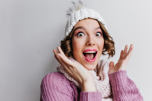Retrato de primer plano de mujer sorprendida de pelo corto con labios rosados posando con sombrero de invierno. sorprendida chica blanca en suéter morado.