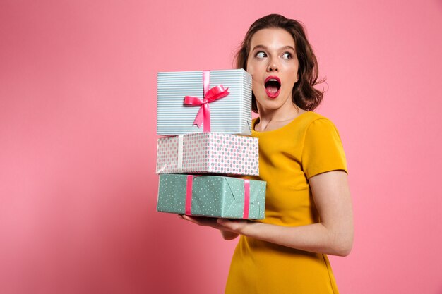 Retrato de primer plano de niña bonita sorprendida con maquillaje brillante con montón de regalos, mirando a un lado