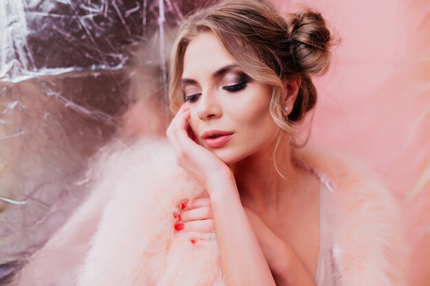 Retrato de primer plano de niña elegante con hermoso maquillaje, posando mientras piensa en sus sueños. Hermosa joven rubia en moda abrigo de piel rosa de pie sobre fondo brillante con los ojos cerrados