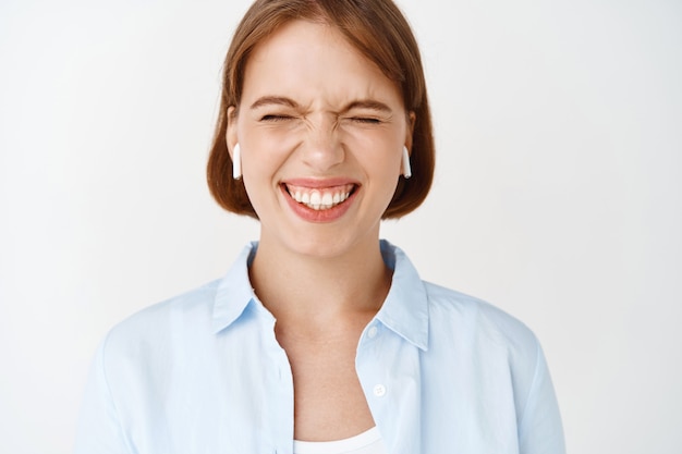 Retrato de primer plano de niña emocionada sonriendo con los ojos cerrados, disfrutando de escuchar música en auriculares inalámbricos, de pie en la pared blanca