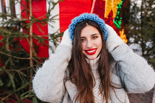 Retrato de primer plano de niña entusiasta con sombrero azul posando con expresión de cara feliz frente a árboles de Navidad. Foto al aire libre de mujer glamorosa con cabello oscuro de pie junto a la decoración de año nuevo.