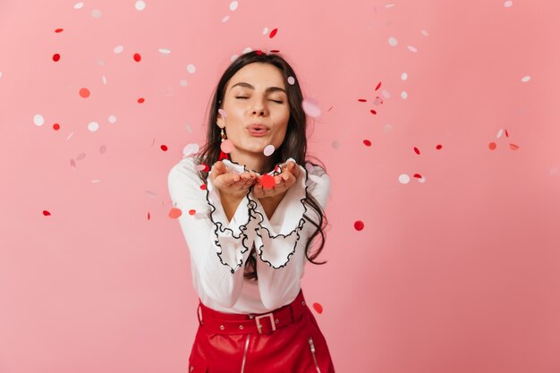 Retrato de primer plano de niña en falda de cuero enviando beso de aire sobre fondo rosa con confeti.