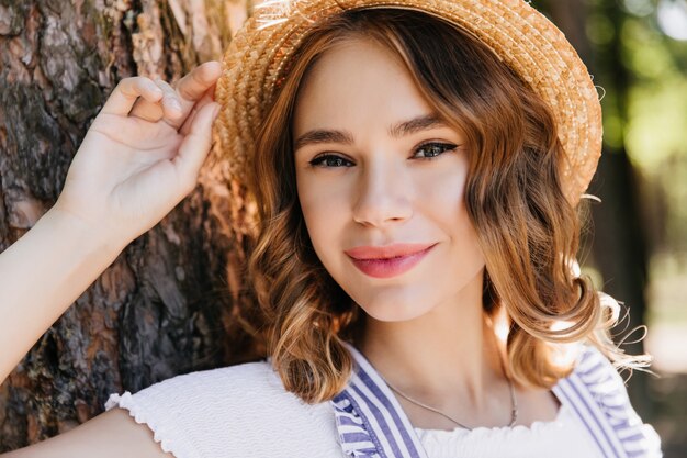 Retrato de primer plano de niña interesada en sombrero de paja posando cerca de árbol. Foto al aire libre de elegante mujer caucásica con ojos azules descansando en el bosque.