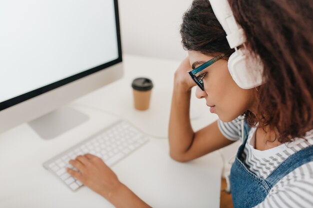 Retrato de primer plano de niña negra cansada en grandes auriculares blancos sentado en el lugar de trabajo y escribiendo en el teclado