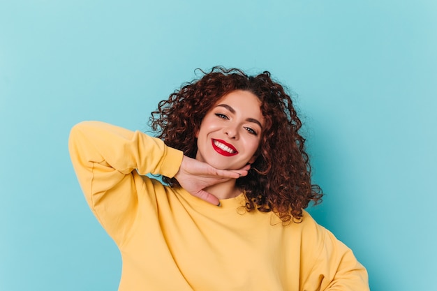 Retrato de primer plano de una niña de ojos azules con labios rojos. Mujer rizada en suéter amarillo toca su barbilla y posa en el espacio azul.