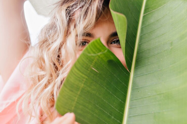 Foto gratuito retrato de primer plano de niña de ojos azules posando juguetonamente con planta. encantadora dama rubia tímida que esconde la cara detrás de la hoja verde.