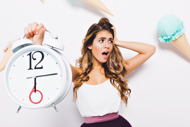 Retrato de primer plano de niña rizada decepcionada mirando a otro lado, con atuendos de moda. Mujer joven con expresión de la cara de pánico sosteniendo un gran reloj de alarma en la pared decorada.