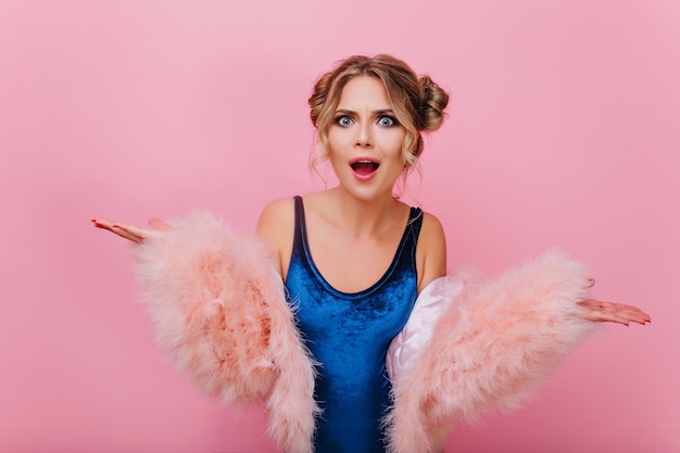 Retrato de primer plano de niña rizada descontenta en traje azul y abrigo mullido, de pie sobre fondo rosa. Mujer joven indignada con lindo peinado posando con las manos saludando y la boca abierta