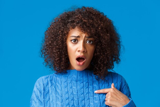 Retrato de primer plano ofendido y molesto tonta tímida mujer afroamericana fue acusada o culpada, señalando a sí misma y luciendo insultada, jadeando mirando algo injusto, de pie pared azul.