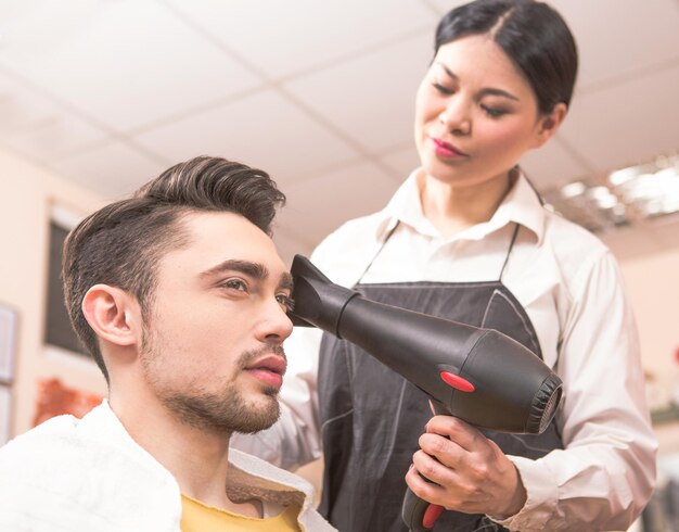 Retrato de primer plano de peluquería secando el cabello de un hombre guapo con secador de pelo en el salón de peluquería. Hombre mirando en el espejo.