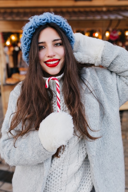 Retrato de primer plano de la romántica chica europea con cabello oscuro posando con dulce piruleta de Navidad. Foto de modelo de mujer bastante caucásica en guantes blancos y sombrero azul divirtiéndose