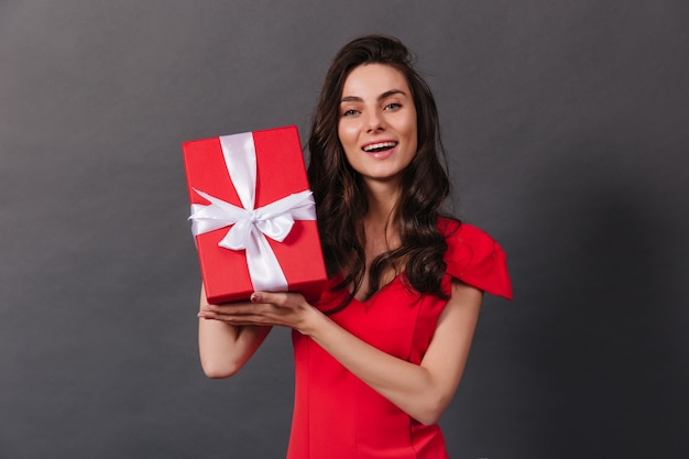 Retrato de primer plano de sonriente mujer morena con caja decorada con regalo. La dama de rojo está sonriendo sinceramente sobre fondo negro.