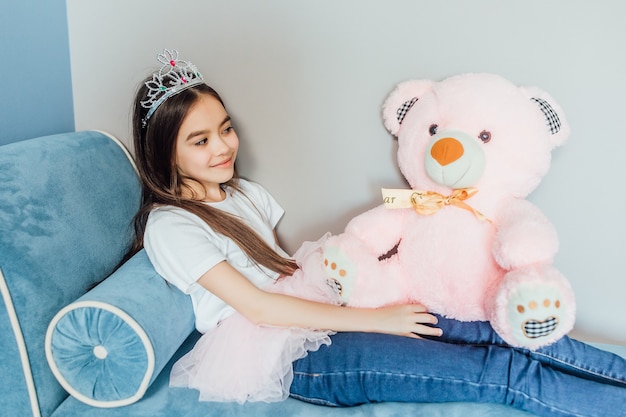 Retrato de princesa feliz jugando con oso rosa y con corona en la cabeza.