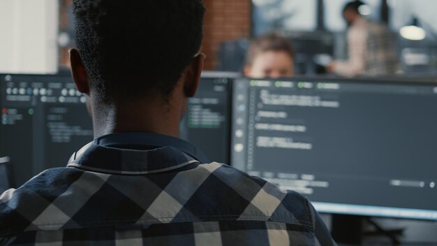 Retrato de un programador afroamericano de computación en la nube en línea en una computadora portátil sentada en el escritorio con múltiples pantallas de código de análisis. Vista sobre el hombro del desarrollador de aplicaciones que trabaja en la interfaz de usuario.