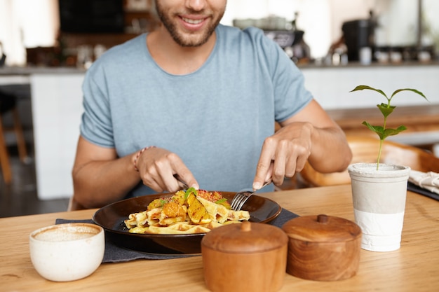 Retrato recortado de feliz joven barbudo en camiseta sonriendo alegremente mientras disfruta de una sabrosa comida durante el almuerzo en el acogedor restaurante, sentado en la mesa de madera