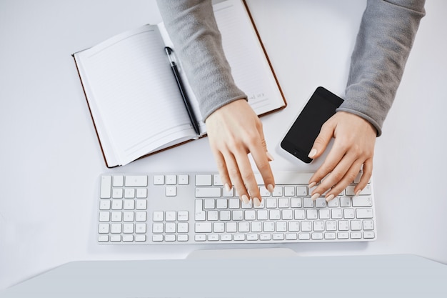 Foto gratuita retrato recortado de manos de mujer escribiendo en el teclado y trabajando con computadoras y gadgets. mujer independiente moderna que diseña un nuevo proyecto para la empresa, tomando notas en un cuaderno y un teléfono inteligente