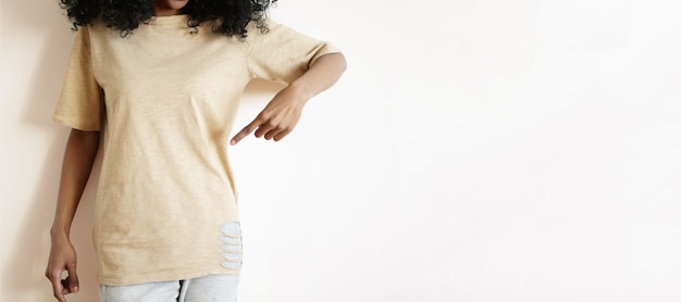 Retrato recortado de modelo de mujer africana joven con ropa casual mostrando su camiseta vacía con el dedo índice