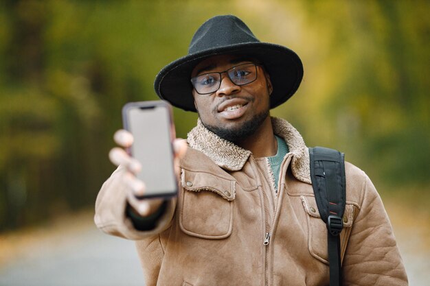 Retrato recortado del viajero masculino negro que muestra el teléfono celular de la pantalla de copia en blanco para su información o publicidad. Enfoque selectivo. Hombre de pie afuera y vestido con una chaqueta marrón, gafas y negro