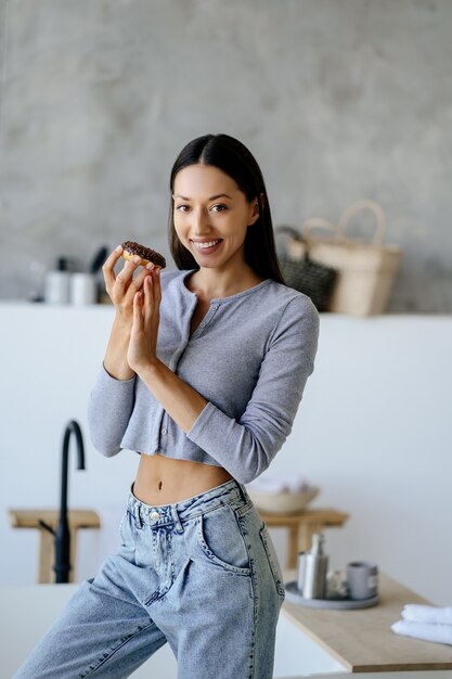 Retrato de regocijo mujer sosteniendo sabroso buñuelo en casa. Concepto de comida poco saludable.