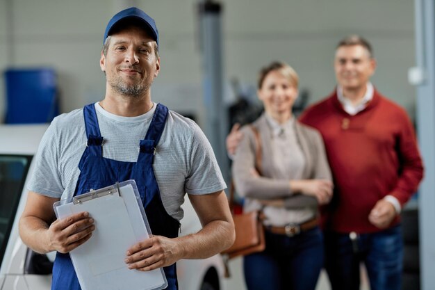 Retrato de un reparador de automóviles feliz mirando la cámara mientras sus clientes están parados en el fondo