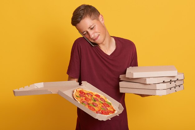 Retrato del repartidor hablando por teléfono, vistiendo una camiseta informal de color burdeos, sosteniendo cajas con pizza, recibe un nuevo pedido a través de su teléfono inteligente