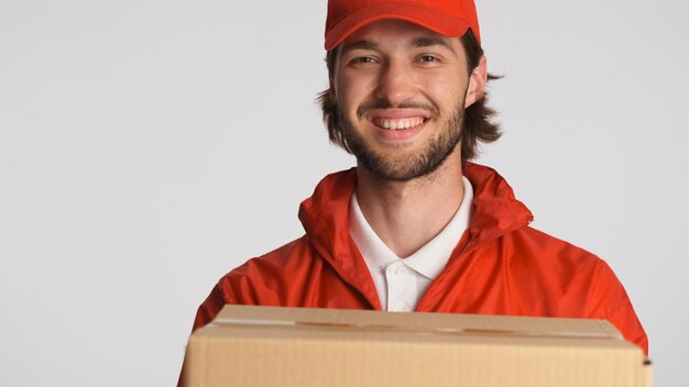 Retrato del repartidor que sostiene el paquete al cliente en la cámara aislada Atractivo mensajero masculino barbudo que parece feliz en el trabajo