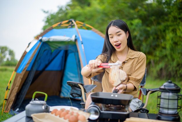 Retrato de Retrato de una joven asiática feliz acampando sola barbacoa de cerdo a la parrilla en la bandeja de picnic y cocinando comida mientras se sienta en una silla en el camping