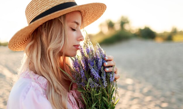 Retrato romántico de cerca o encantadora chica rubia con sombrero de paja huele flores en la playa por la noche, cálidos colores del atardecer. Ramo de lavanda. Detalles.