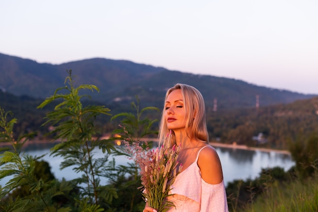 Retrato romántico de joven mujer caucásica en vestido de verano disfrutando de relajarse en el parque en la montaña con increíbles vistas al mar tropical