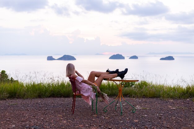 Retrato romántico de joven mujer caucásica en vestido de verano disfrutando de relajarse en el parque en la montaña con increíbles vistas al mar tropical