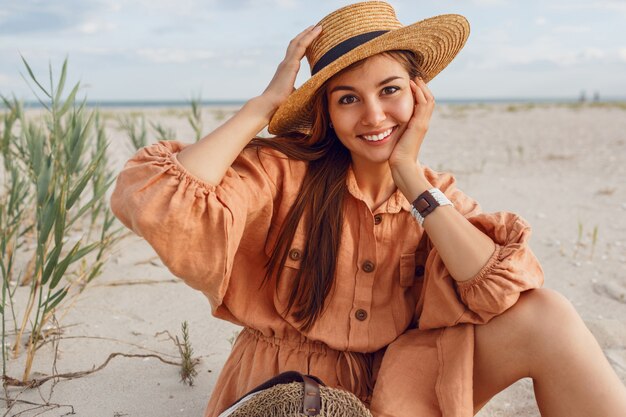 Retrato romántico de mujer sonriente con sombrero de paja y elegante vestido de lino. Chica soñadora escalofriante cerca del océano. Tendencia de la moda de verano.