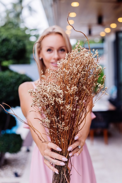 Retrato romántico de mujer en vestido lindo de noche rosa mantenga flores silvestres fuera de villa tropical de lujo Hermosa mujer con ramo