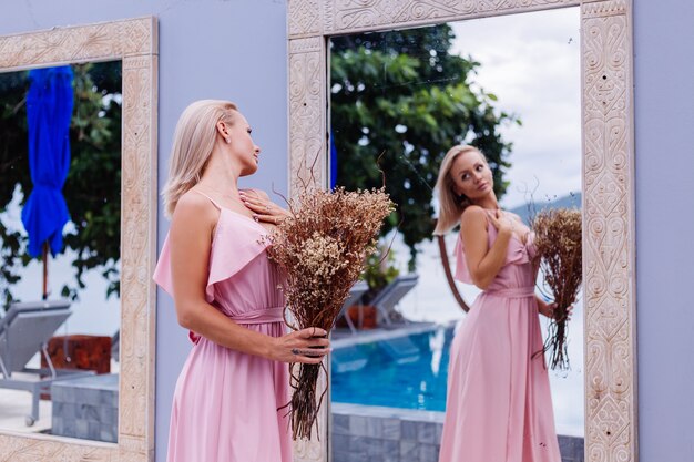 Retrato romántico de mujer en vestido lindo de noche rosa mantenga flores silvestres fuera de villa tropical de lujo Hermosa mujer con ramo