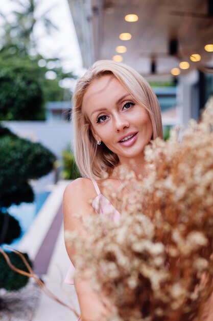 Retrato romántico de mujer en vestido lindo de noche rosa mantenga flores silvestres fuera de villa tropical de lujo Hermosa mujer con ramo