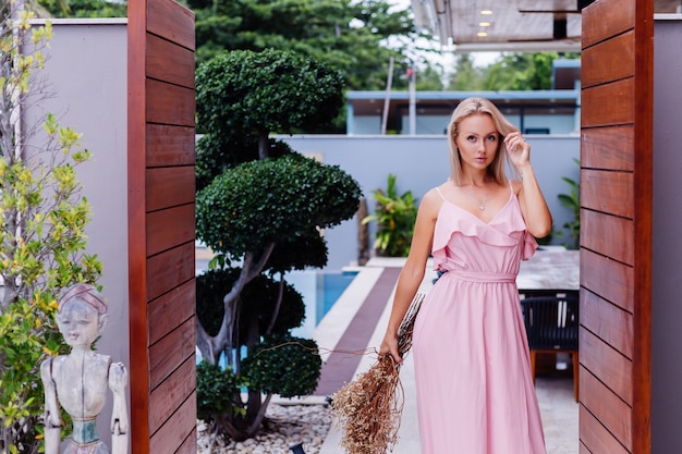 Foto gratuita retrato romántico de mujer en vestido lindo de noche rosa mantenga flores silvestres fuera de villa tropical de lujo hermosa mujer con ramo