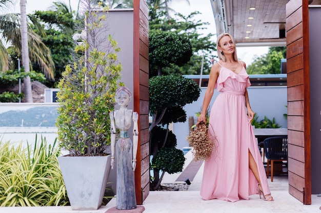 Retrato romántico de mujer en vestido lindo de noche rosa mantenga flores silvestres fuera de villa tropical de lujo Hermosa mujer con ramo