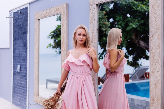 Retrato romántico de mujer en vestido lindo de noche rosa mantenga flores silvestres fuera de villa tropical de lujo Hermosa mujer con ramo