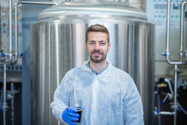 Foto gratuita retrato de rubia tecnólogo sosteniendo un vaso de producto de bebida en la línea de producción de cerveza de fábrica