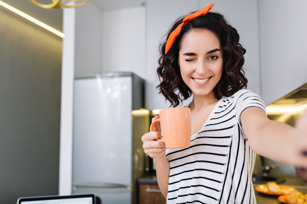 Retrato de selfie doméstico con estilo de mujer joven y bonita sonriendo, divirtiéndose con una taza de té. Fines de semana en casa, apartamento moderno, ocio, comodidad.