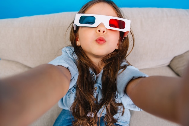 Foto gratuita retrato de selfie con estilo de niña encantadora en gafas 3d enviando un beso a la cámara. relajarse en el sofá sobre fondo azul, vistiendo ropa de jeans, cabello largo morena, expresando felicidad